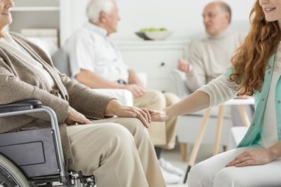 Man in a Wheelchair in a Nursing Home Holding Onto Younger Woman's Hand on His Knee